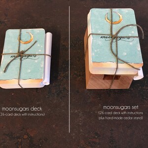 May include: Two versions of a moonsugars deck of cards. The deck on the left is wrapped in twine and includes instructions. The deck on the right is on a wooden stand and includes instructions. Both decks feature a crescent moon and the word "moonsugars" on the front.