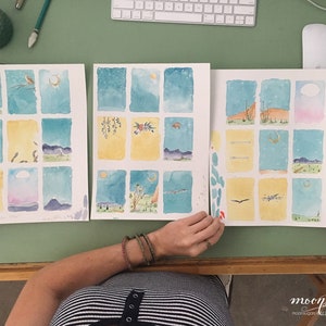 May include: Three watercolor paintings on paper, each divided into nine squares. The paintings depict a night sky with stars and a crescent moon, with different scenes in each square. The paintings are on a green table with a person's hand holding the rightmost painting.