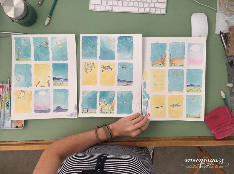 May include: Three watercolor paintings on paper, each divided into nine squares. The paintings depict a night sky with stars and a crescent moon, with different scenes in each square. The paintings are on a green table with a person's hand holding the rightmost painting.