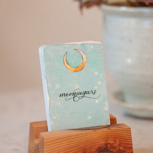May include: A light blue card with a crescent moon and the text "moonsugars" in black cursive. The card is sitting in a wooden stand.