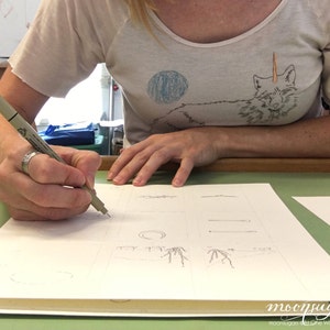 May include: A person is drawing on a white sheet of paper with a black pen. The paper is divided into squares, and each square contains a different drawing. The person is wearing a white t-shirt with a fox design on it.