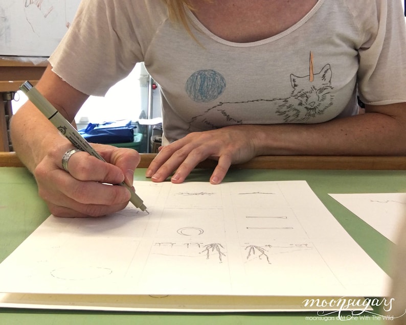May include: A person is drawing on a white sheet of paper with a black pen. The paper is divided into squares, and each square contains a different drawing. The person is wearing a white t-shirt with a fox design on it.
