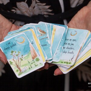 May include: A set of watercolor illustrated affirmation cards with a blue, green, and yellow color scheme. The cards feature a crescent moon, a white owl, and a mountain. The text on the cards reads "I am always guided." and "Close my eyes, relax my jaw, my shoulders, deep breaths."