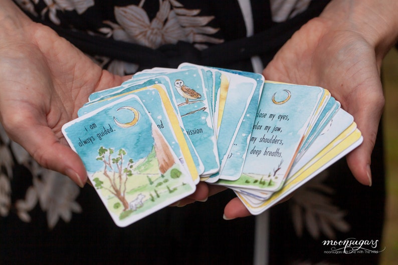 May include: A set of watercolor illustrated affirmation cards with a blue, green, and yellow color scheme. The cards feature a crescent moon, a white owl, and a mountain. The text on the cards reads "I am always guided." and "Close my eyes, relax my jaw, my shoulders, deep breaths."