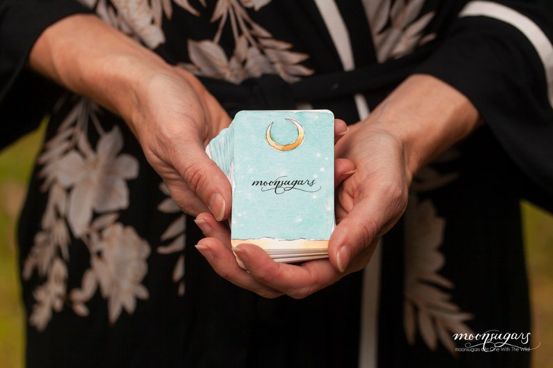 May include: A person is holding a deck of blue and gold moon oracle cards. The cards have a crescent moon and the word "moonjaguars" printed on them.