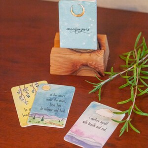 May include: Three watercolor cards with inspirational quotes about self-care and healing. The cards feature a crescent moon, a mountain range with a winding path, and a cloud in a blue sky. The cards are part of a deck called "moonsugars".