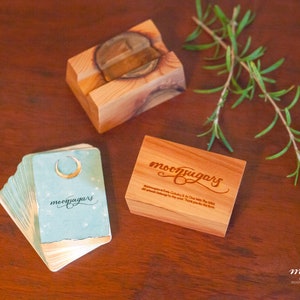 May include: A set of blue and gold oracle cards with a crescent moon design and the text "moonpugars" on the front. The cards are stacked on a wooden table next to a wooden box with the text "moonpugars" engraved on the lid. A sprig of rosemary is also on the table.