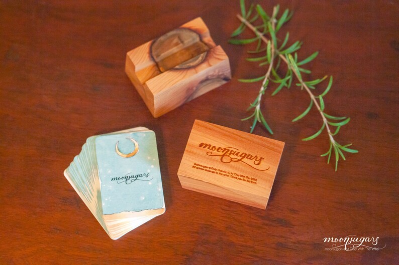 May include: A set of blue and gold oracle cards with a crescent moon design and the text "moonpugars" on the front. The cards are stacked on a wooden table next to a wooden box with the text "moonpugars" engraved on the lid. A sprig of rosemary is also on the table.