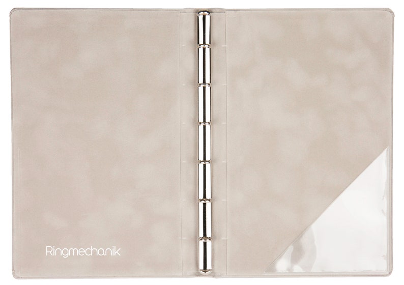 May include: A beige leather binder with a clear plastic pocket on the right side. The binder has a silver ring mechanism and the text "Ringmechanik" is printed on the front cover.