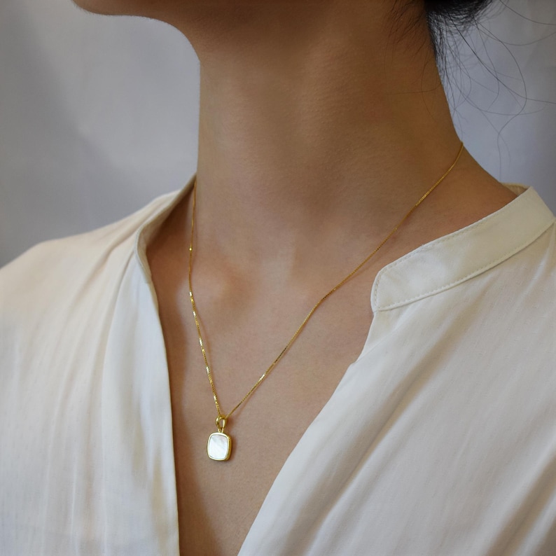 May include: A gold chain necklace with a square-shaped pendant featuring a white, iridescent stone.