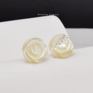 Op de afbeelding: Een paar witte parelmoer rozen oorringen. De oorringen zijn gevormd als rozen en hebben een delicate, vrouwelijke uitstraling.