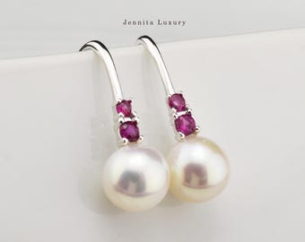 May include: A pair of silver drop earrings featuring two small round pink stones set vertically above a single white pearl. The metal hooks curve upward, and the text Jennita Luxury is printed in black above the jewelry pieces.