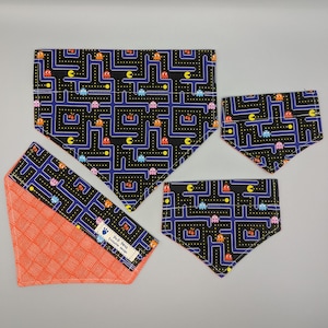 May include: Four different sizes of black and blue fabric dog bandanas with a Pac-Man pattern. The bandanas have a white and orange border. The bandanas are on a white background.