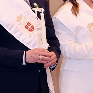 May include: Two people wearing white stoles embroidered with a coat of arms, holding a lit candle. The coat of arms features a crown and a shield with a cross design. One person wears a dark suit, the other a white jacket.
