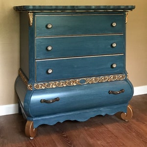 May include: A blue chest of drawers with gold accents and ornate carvings. The chest has four drawers with crystal knobs and a curved bottom.