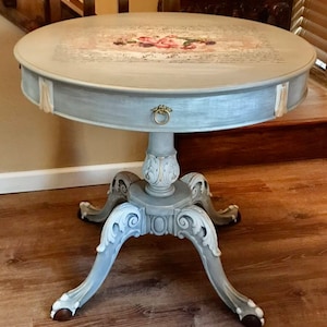May include: A round, gray, wooden side table with a single drawer and ornate legs. The top of the table has a decorative floral design and a handwritten inscription.