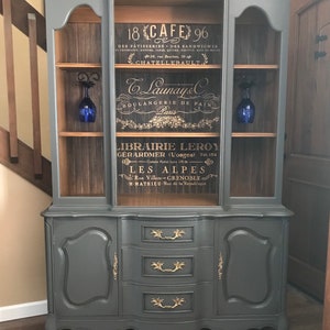 May include: A gray painted antique hutch with glass doors and three drawers. The hutch has a wood-grain back panel with gold lettering that reads "18 CAFE 96", "Launay & C Boulangerie de Pain Paris", "LIBRAIRIE LEROY GERARDMER (Vosges) Tel. 154", "LES ALPES Rue Villars - GRENOBLE - M-MATHIEU - Rue de la Republique".