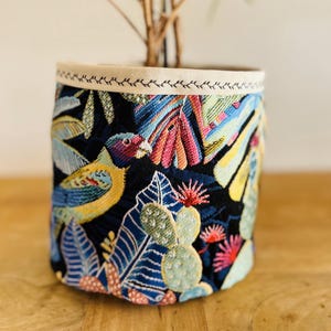 Può includere: Un vaso da fiori in tessuto nero con una stampa tropicale colorata che presenta uccelli, foglie e fiori. Il vaso ha un bordo bianco attorno alla parte superiore.