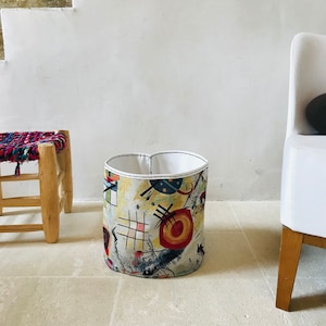 May include: A cylindrical fabric storage bin with a white top and a colorful abstract design featuring red, yellow, black, and blue. The bin is sitting on a light beige tiled floor.