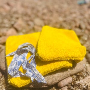 May include: A bright yellow terry cloth pouch featuring a light blue and white floral patterned ribbon tie. The rectangular fabric accessory has a textured surface and is folded over itself, showcasing the vibrant yellow color and soft material.