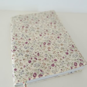 Peut inclure: Un journal à couverture crème orné d'un motif floral et d'un signet en ruban assorti. La couverture présente un motif répété de petites fleurs violettes, bleues et crème. Le journal est ouvert, révélant des pages blanches.
