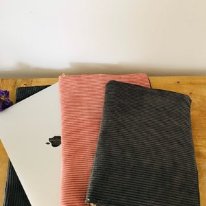 May include: Three laptop sleeves, one black, one pink, and one gray, are stacked on top of each other. The sleeves are made of corduroy fabric and have a zipper closure.