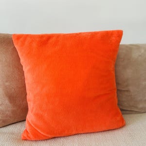 May include: A square, vibrant orange cushion with a soft, fuzzy texture. The cushion is the main focus, set against a neutral backdrop with a glimpse of a brown cushion on the left. The cushion is on a light-coloured sofa.