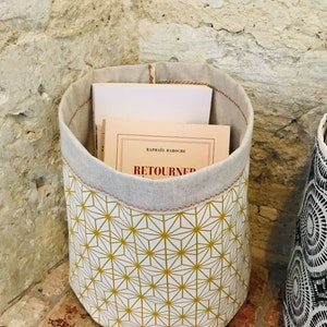 Op de afbeelding: Een witte stoffen opbergmand met een geel geometrisch patroon. De mand heeft een denim bovenkant en is gevuld met boeken. De tekst "RETOURNER" is zichtbaar op het boek.