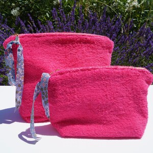 Pode incluir: Duas bolsas de zíper de felpa rosa com puxadores de fita floral azul e branca. A bolsa maior está atrás da menor.