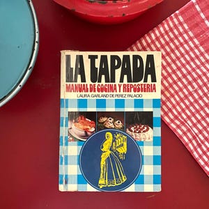 May include: A vintage cookbook titled "LA TAPADA MANUAL DE COCINA Y REPOSTERIA" with a blue and white checkered pattern. The cover features illustrations of food and a woman in a yellow dress. Red and blue enamelware and a red and white checkered cloth are in the background.