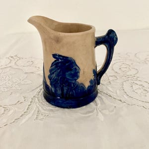 May include: Small, vintage ceramic pitcher with a tan body and a dark blue handle and base. The pitcher features a blue relief design of a Native American profile. The spout is curved, and the handle is ornate.