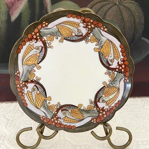 May include: A decorative plate with a scalloped edge, featuring a design of fish and berries. The plate has a white center, a dark green border, and is displayed on a gold-colored metal stand. The fish are gray and gold.