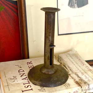 May include: A vintage, antique candle holder made of aged metal. The candle holder has a circular base, a tall, slender stem, and a small, flat top. It is resting on a stack of books with the title "The Island at the Center of the World."