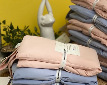 May include: Stacks of pink and blue rectangular pillows, tied with white ribbon, are displayed. The pillows have a tag that reads "The Sankalpa Project". A white frog statue is in the background, with a yellow wall.