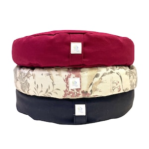 Puede incluir: Tres cojines de meditación redondos apilados uno encima del otro. El cojín superior es de color rojo oscuro, el cojín del medio es de color beige con un patrón floral y el cojín inferior es negro. Cada cojín tiene una pequeña etiqueta redonda cosida en el lateral.