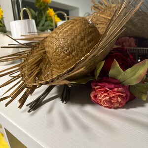 Puede incluir: Un sombrero de paja tejido con una corona redonda y ala ancha, adornado con flecos de paja seca. El sombrero se combina con un ramo de rosas rojas y rosas y hojas verdes, creando una est&eacute;tica r&uacute;stica y natural.