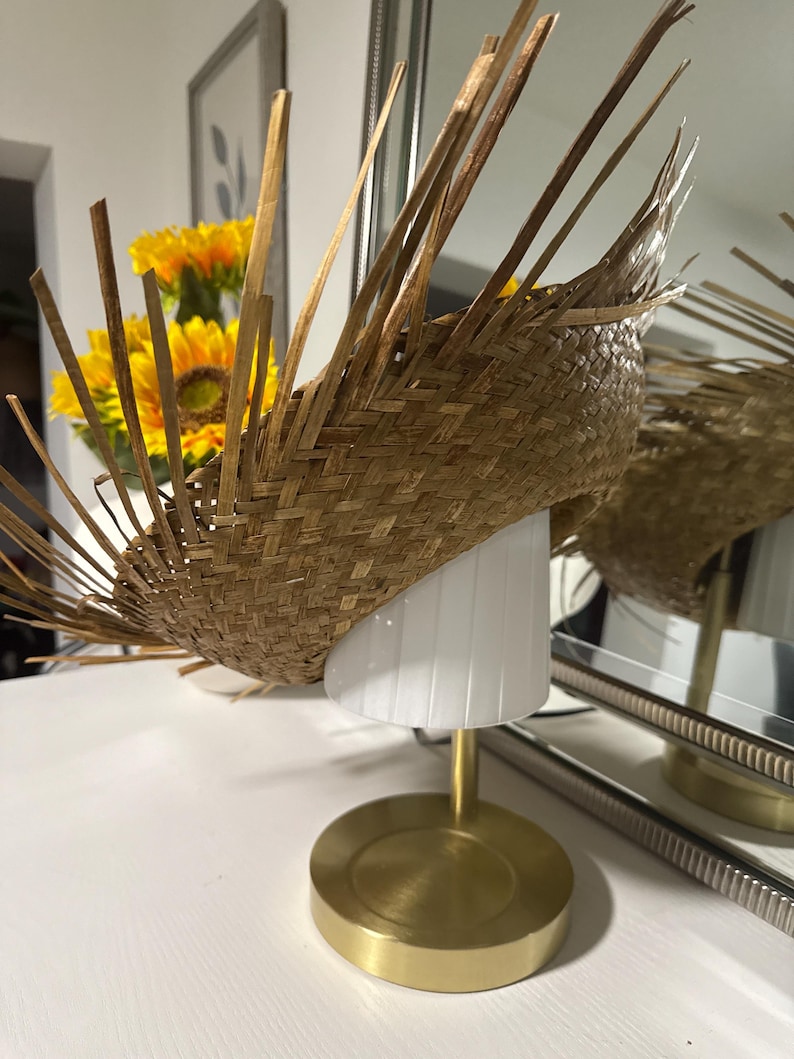 Puede incluir: Un sombrero de paja tejido con un dise&ntilde;o &uacute;nico, con un ala ancha y un tejido texturizado. El sombrero se exhibe sobre una l&aacute;mpara blanca con una base dorada, creando un contraste llamativo. Se destaca el color y la textura natural del sombrero.