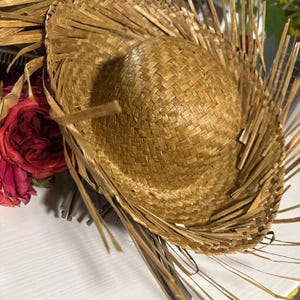 Puede incluir: Un sombrero de paja con una corona redonda y un ala ancha. El sombrero est&aacute; hecho de paja tejida y tiene flecos decorativos de paja alrededor del ala. El sombrero es de color marr&oacute;n claro. El sombrero est&aacute; junto a un ramo de rosas rojas.