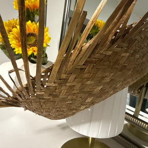 Puede incluir: Una cesta de bamb&uacute; tejida con un dise&ntilde;o en forma de abanico. La cesta es de color marr&oacute;n claro y tiene una superficie texturizada. La cesta se exhibe cerca de una l&aacute;mpara blanca y girasoles amarillos.