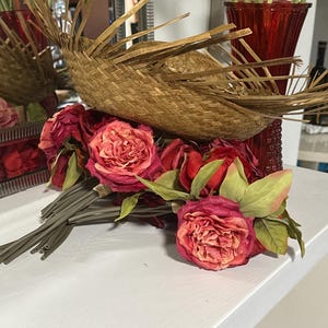 Puede incluir: Un ramo de rosas rojas con hojas y tallos verdes descansa sobre una superficie blanca. Una cesta tejida con un dise&ntilde;o en forma de abanico se encuentra encima de las rosas. Un jarr&oacute;n rojo es visible en el fondo.