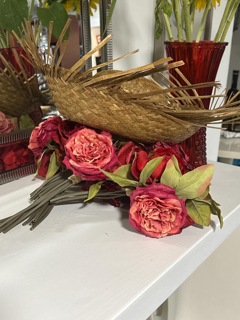 Puede incluir: Un ramo de rosas rojas con hojas y tallos verdes descansa sobre una superficie blanca. Una cesta tejida con un dise&ntilde;o en forma de abanico se encuentra encima de las rosas. Un jarr&oacute;n rojo es visible en el fondo.