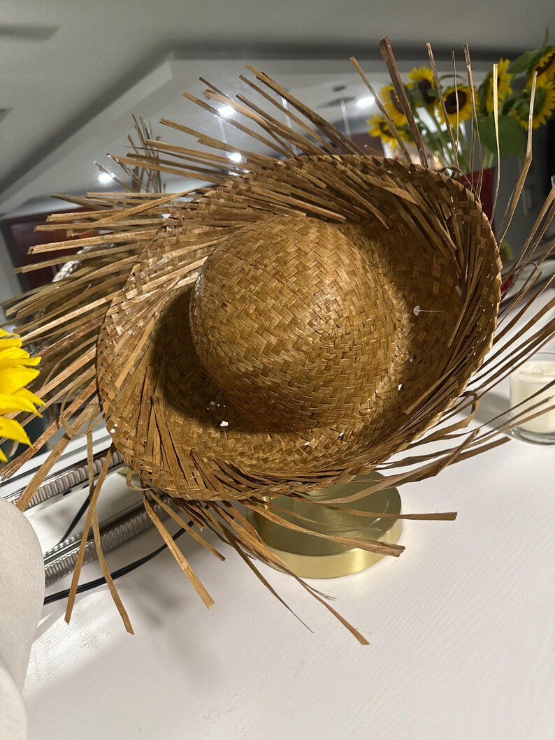 Puede incluir: Un sombrero de paja tejido con una corona redondeada y un ala ancha, adornado con flecos de paja radiantes. El sombrero es de color marr&oacute;n natural, lo que sugiere un tema r&uacute;stico o tropical. El sombrero est&aacute; sobre una base dorada.