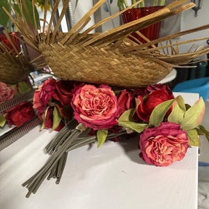 Puede incluir: Una cesta tejida con tiras de bamb&uacute; radiantes se asienta sobre un ramo de rosas rojas y rosas. Las rosas tienen hojas verdes y tallos grises. El fondo incluye girasoles y un jarr&oacute;n rojo.