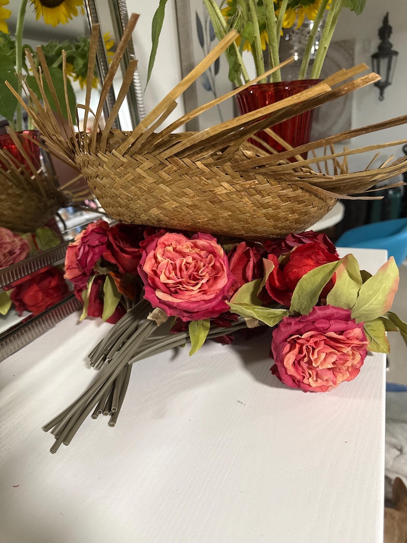 Puede incluir: Una cesta tejida con tiras de bamb&uacute; radiantes se asienta sobre un ramo de rosas rojas y rosas. Las rosas tienen hojas verdes y tallos grises. El fondo incluye girasoles y un jarr&oacute;n rojo.