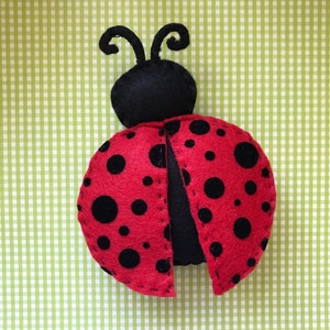 Puede incluir: Una mariquita roja con manchas negras y antenas negras. La mariquita está hecha de fieltro y tiene una forma tridimensional.