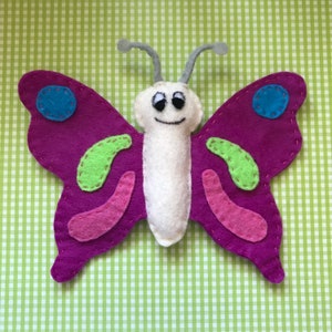 May include: A purple felt butterfly with green, pink, and blue accents. The butterfly has a white felt body with a smiling face.
