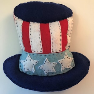 Könnte beinhalten: Ein patriotischer Filzhut in Form eines Zylinders mit roten und weißen Streifen und drei weißen Sternen auf blauem Hintergrund.