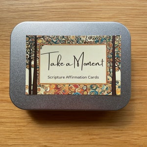 May include: A silver tin with a colourful design featuring a tree motif. The tin has the text "Take a Moment" and "Scripture Affirmation Cards" printed on it.
