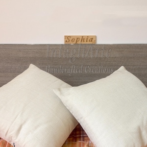 Può includere: Due cuscini quadrati bianchi sporchi con una superficie strutturata poggiano su una coperta fantasia. Un cartello di legno sopra i cuscini recita "Sophia". Lo sfondo presenta una testiera in legno con il testo "IntagliArte Handcrafted Creations".