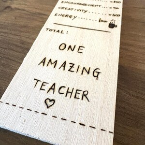 Può includere: Una targa di legno a forma di ricevuta, con le parole "TEACHER RECEIPT" in alto. Elenca qualità come "PATIENCE" e "KINDNESS", e si conclude con "ONE AMAZING TEACHER" e un simbolo a forma di cuore. Il testo è inciso nel legno.